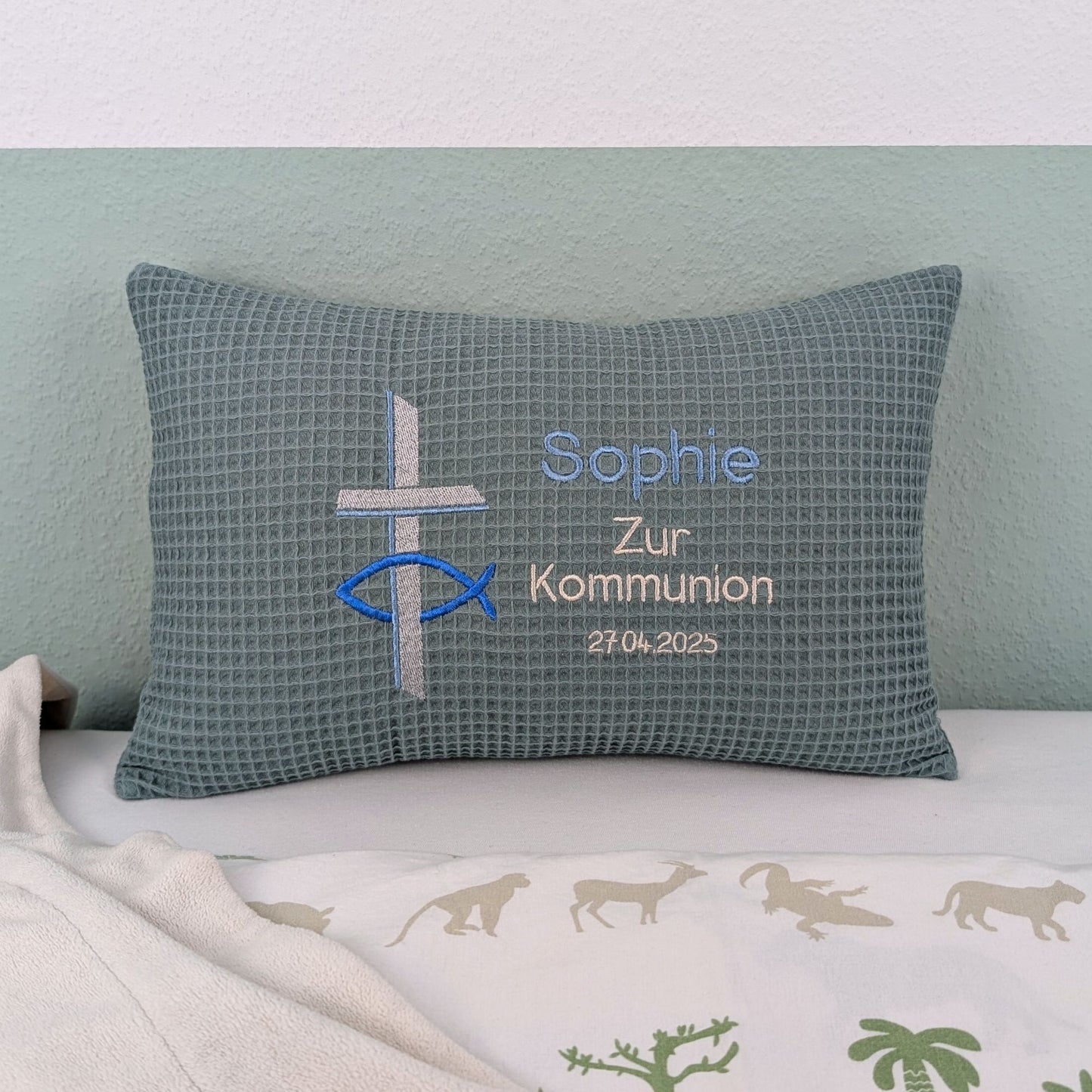 Rechteckiges Kissen in türkiser Farbe mit Waffelstruktur liegt auf einem Bett. Auf dem Kissen sind ein gesticktes Kreuz und ein christliches Ichthys-Symbol (Fisch) sowie der Text „Sophie – Zur Kommunion – 27.4.2025“ in verschiedenen Farben eingestickt. Das Kissen dient als personalisiertes Geschenk zur Erstkommunion.