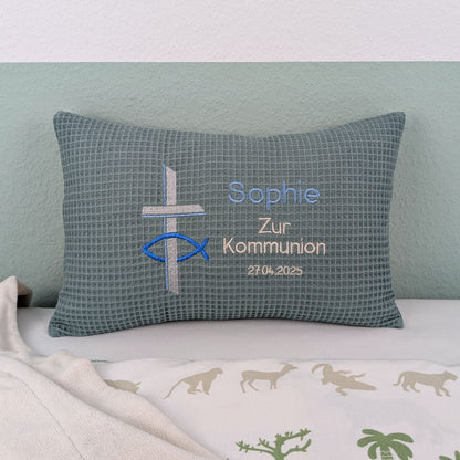 Rechteckiges Kissen in türkiser Farbe mit Waffelstruktur liegt auf einem Bett. Auf dem Kissen sind ein gesticktes Kreuz und ein christliches Ichthys-Symbol (Fisch) sowie der Text „Sophie – Zur Kommunion – 27.4.2025“ in verschiedenen Farben eingestickt. Das Kissen dient als personalisiertes Geschenk zur Erstkommunion.