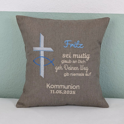 "Besticktes Kissen zur Kommunion. Das Kissen ist grau und zeigt eine Stickerei mit einem christlichen Kreuz und einem Fischsymbol in Blau und Silber. Der Name 'Fritz' ist in blauer Schrift gestickt, gefolgt von einem ermutigenden Spruch in Beige: 'Sei mutig, glaub an Dich, geh Deinen Weg, gib niemals auf'. Darunter steht 'Kommunion 11.05.2025' in weißer Schrift. Das Kissen liegt auf einem weißen, Untergrund."