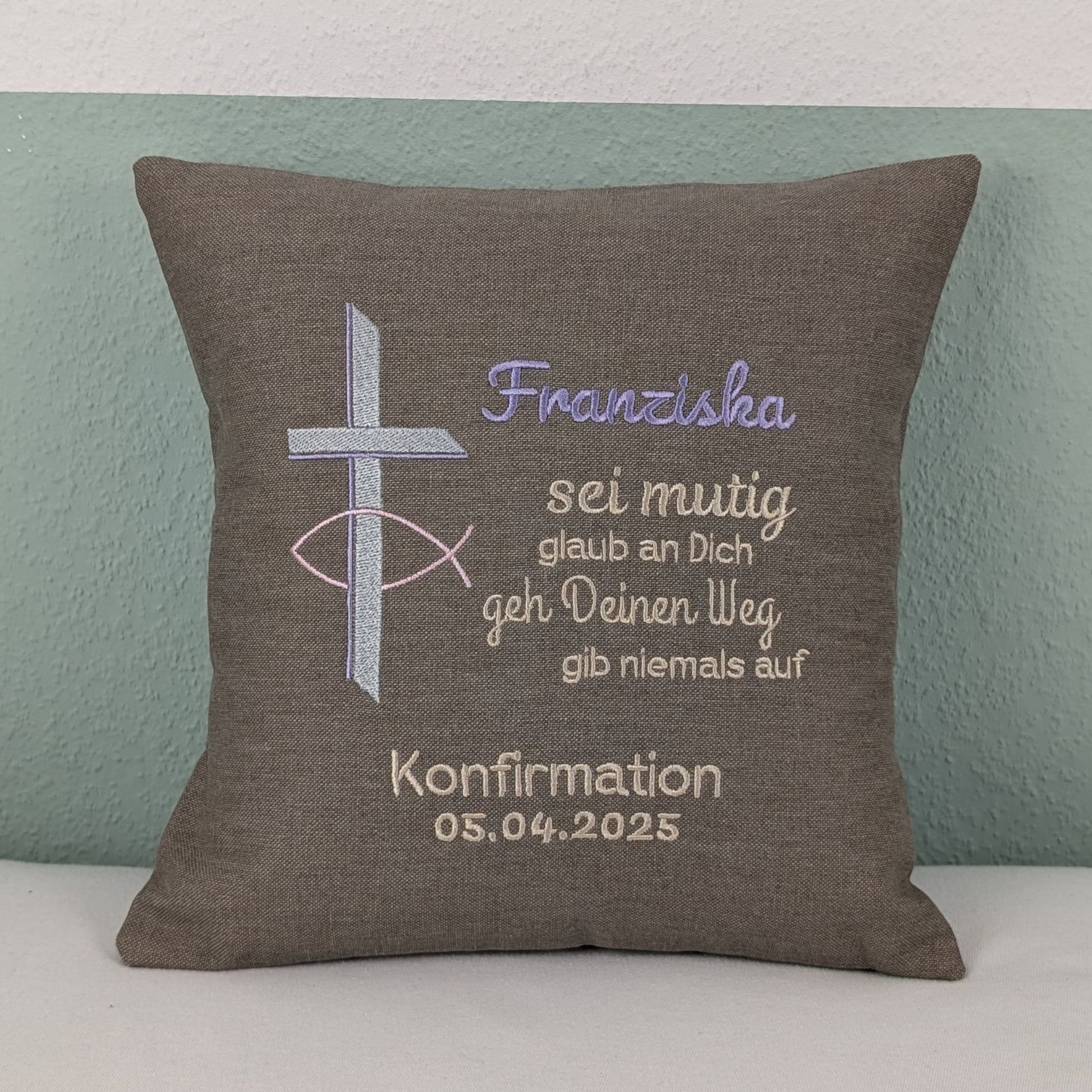 "Besticktes Kissen zur Konfirmation. Das Kissen ist grau und zeigt eine Stickerei mit einem christlichen Kreuz und einem Fischsymbol in lavendel und Silber. Der Name 'Noah' ist in blauer Schrift gestickt, gefolgt von einem ermutigenden Spruch in Beige: 'Sei mutig, glaub an Dich, geh Deinen Weg, gib niemals auf'. Darunter steht 'Konfirmation 05.04.2025' in weißer Schrift. Das Kissen liegt auf einem weißen, Untergrund."