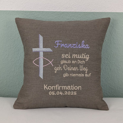 "Besticktes Kissen zur Konfirmation. Das Kissen ist grau und zeigt eine Stickerei mit einem christlichen Kreuz und einem Fischsymbol in lavendel und Silber. Der Name 'Noah' ist in blauer Schrift gestickt, gefolgt von einem ermutigenden Spruch in Beige: 'Sei mutig, glaub an Dich, geh Deinen Weg, gib niemals auf'. Darunter steht 'Konfirmation 05.04.2025' in weißer Schrift. Das Kissen liegt auf einem weißen, Untergrund."