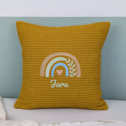 Kissen in der Größe 30 x 30 cm, aus Waffelstoff in der Farbe ocker, bestickt mit einem Regenbogen und darunter steht mittig eine Name.