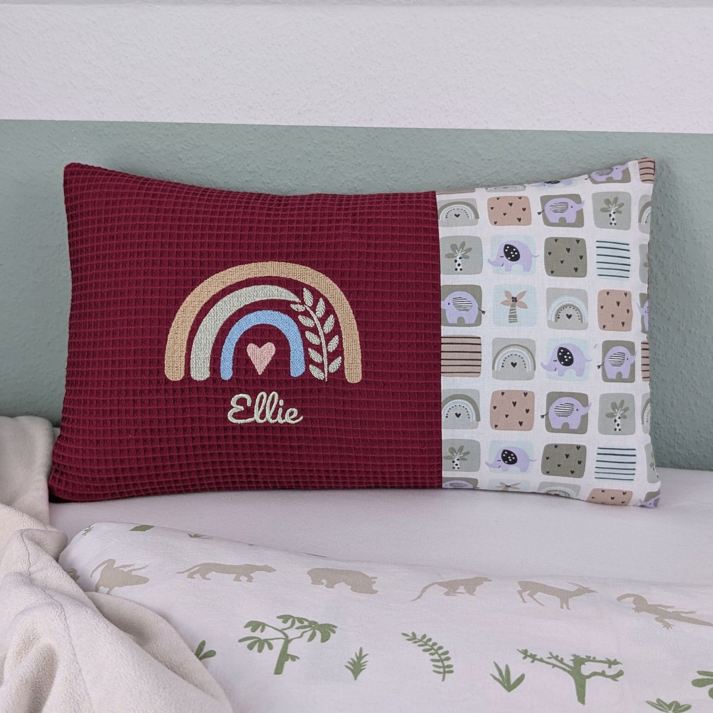 Dekoratives Namenskissen mit gesticktem Namen und Geburtsdaten. Das Kissen hat eine beerefarbene Waffelstoff-Seite mit einer Stickerei eines Regenbogens in Beige, Blau und Gold, ergänzt durch ein Herz- und Ährenmotiv. Darunter steht, der Name vom Kind in grüner Schrift. Die rechte Seite besteht aus einem gemusterten Stoff. Das Kissen liegt auf einer hellen Oberfläche, an eine grün-weiße Wand gelehnt."