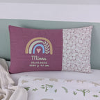 Dekoratives Geburtskissen mit gesticktem Namen und Geburtsdaten. Das Kissen hat eine alt rosa Waffelstoff-Seite mit einer Stickerei eines Regenbogens in Beige, Blau und Gold, ergänzt durch ein Herz- und Ährenmotiv. Darunter steht, der Name vom Kind, sowie Geburtsdatum, Größe und Gewicht in grüner Schrift. Die rechte Seite besteht aus einem gemusterten Stoff. Das Kissen liegt auf einer hellen Oberfläche, an eine grün-weiße Wand gelehnt."