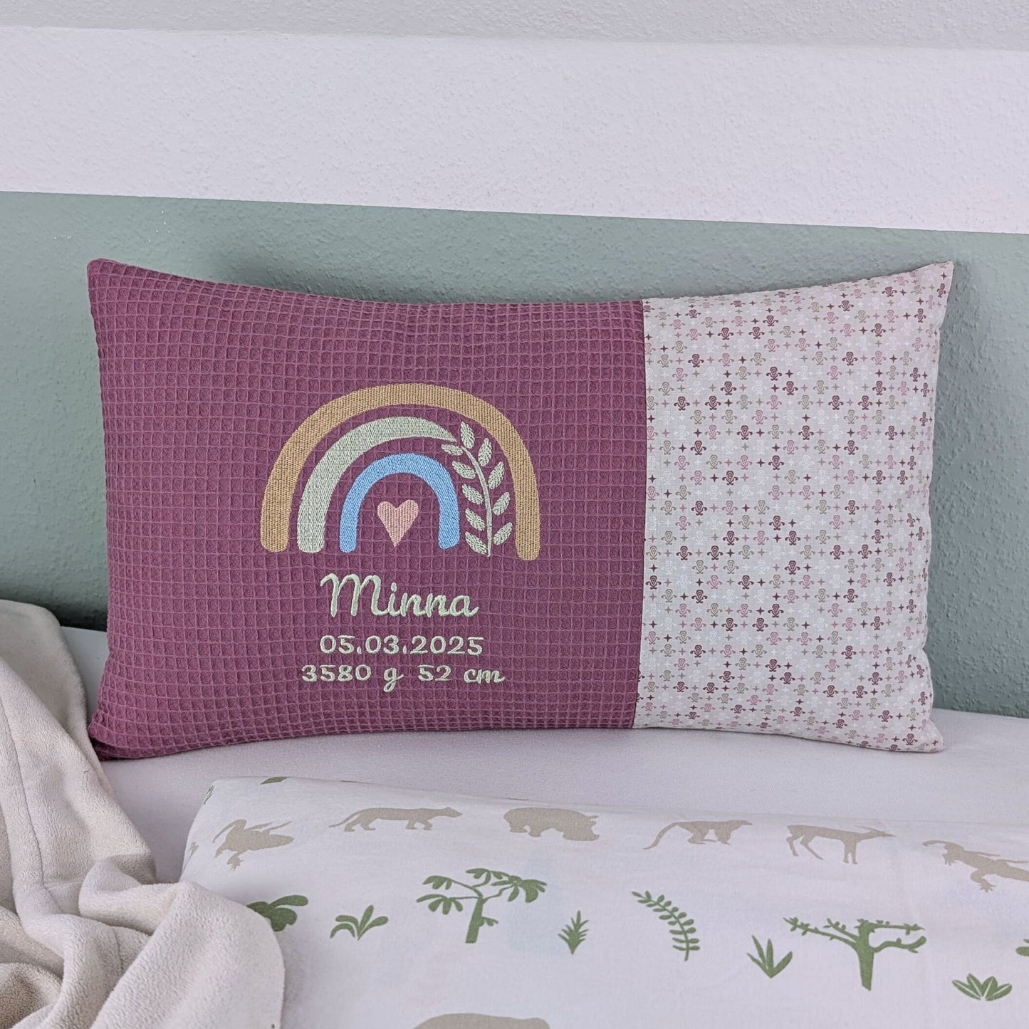 Dekoratives Geburtskissen mit gesticktem Namen und Geburtsdaten. Das Kissen hat eine alt rosa Waffelstoff-Seite mit einer Stickerei eines Regenbogens in Beige, Blau und Gold, ergänzt durch ein Herz- und Ährenmotiv. Darunter steht, der Name vom Kind, sowie Geburtsdatum, Größe und Gewicht in grüner Schrift. Die rechte Seite besteht aus einem gemusterten Stoff. Das Kissen liegt auf einer hellen Oberfläche, an eine grün-weiße Wand gelehnt."