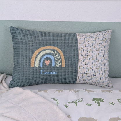 Dekoratives Namenskissen mit gesticktem Namen. Das Kissen hat eine türkisen Waffelstoff-Seite mit einer Stickerei eines Regenbogens in Beige, Blau und Gold, ergänzt durch ein Herz- und Ährenmotiv. Darunter steht, der Name vom Kind in grüner Schrift. Die rechte Seite besteht aus einem gemusterten Stoff. Das Kissen liegt auf einer hellen Oberfläche, an eine grün-weiße Wand gelehnt."