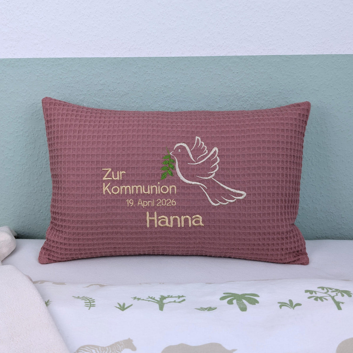 Rechteckiges, altrosa farbendes Kissen aus Waffelstoff liegt auf einem Bett. Auf dem Kissen ist eine weiße Taube mit einem grünen Olivenzweig im Schnabel sowie der gestickte Text „Zur Kommunion am 11. Mai 2025 – und der Name Valentina" zu sehen. Das Kissen dient als persönliches Geschenk zur Erstkommunion.