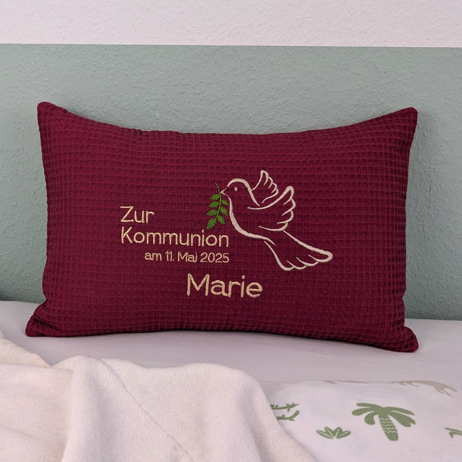 Rechteckiges, beerefarbendes Kissen aus Waffelstoff liegt auf einem Bett. Auf dem Kissen ist eine weiße Taube mit einem grünen Olivenzweig im Schnabel sowie der gestickte Text „Zur Kommunion am 11. Mai 2025 – und der Name Marie" zu sehen. Das Kissen dient als persönliches Geschenk zur Erstkommunion.