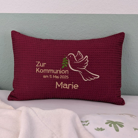 Rechteckiges, beerefarbendes Kissen aus Waffelstoff liegt auf einem Bett. Auf dem Kissen ist eine weiße Taube mit einem grünen Olivenzweig im Schnabel sowie der gestickte Text „Zur Kommunion am 11. Mai 2025 – und der Name Marie" zu sehen. Das Kissen dient als persönliches Geschenk zur Erstkommunion.