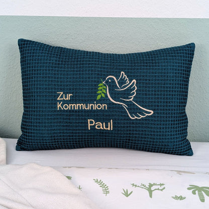 Rechteckiges, petrolfarbendes Kissen aus Waffelstoff liegt auf einem Bett. Auf dem Kissen ist eine weiße Taube mit einem grünen Olivenzweig im Schnabel sowie der gestickte Text „Zur Kommunion – und der Name Paul" zu sehen. Das Kissen dient als persönliches Geschenk zur Erstkommunion.