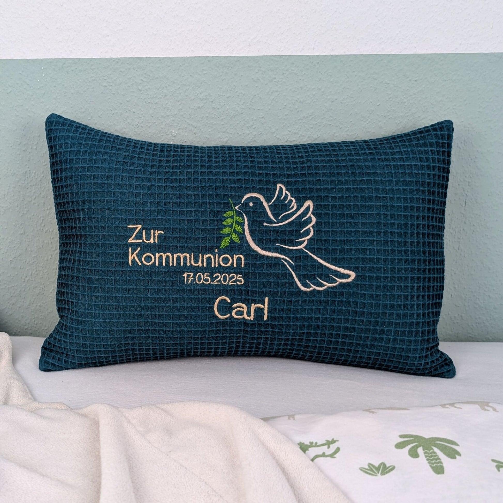 Rechteckiges, petrolfarbendes Kissen aus Waffelstoff liegt auf einem Bett. Auf dem Kissen ist eine weiße Taube mit einem grünen Olivenzweig im Schnabel sowie der gestickte Text „Zur Kommunion 17.05. 2025 – und der Name Carl" zu sehen. Das Kissen dient als persönliches Geschenk zur Erstkommunion.