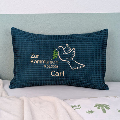 Rechteckiges, petrolfarbendes Kissen aus Waffelstoff liegt auf einem Bett. Auf dem Kissen ist eine weiße Taube mit einem grünen Olivenzweig im Schnabel sowie der gestickte Text „Zur Kommunion 17.05. 2025 – und der Name Carl" zu sehen. Das Kissen dient als persönliches Geschenk zur Erstkommunion.